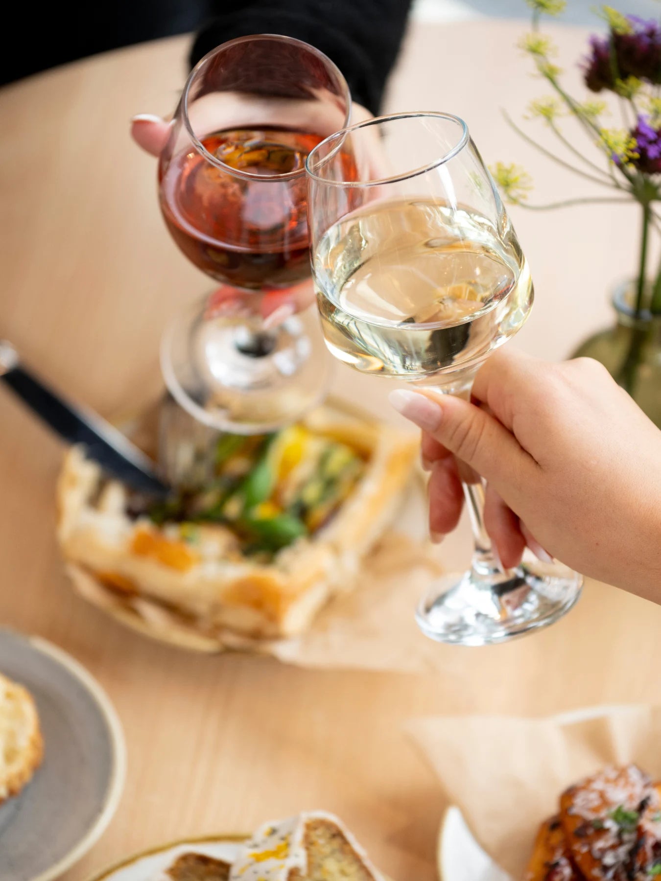 Two hands holding wine glasses at a dining table, with one glass containing white wine and the other containing red wine, with food and flowers blurred in the background.