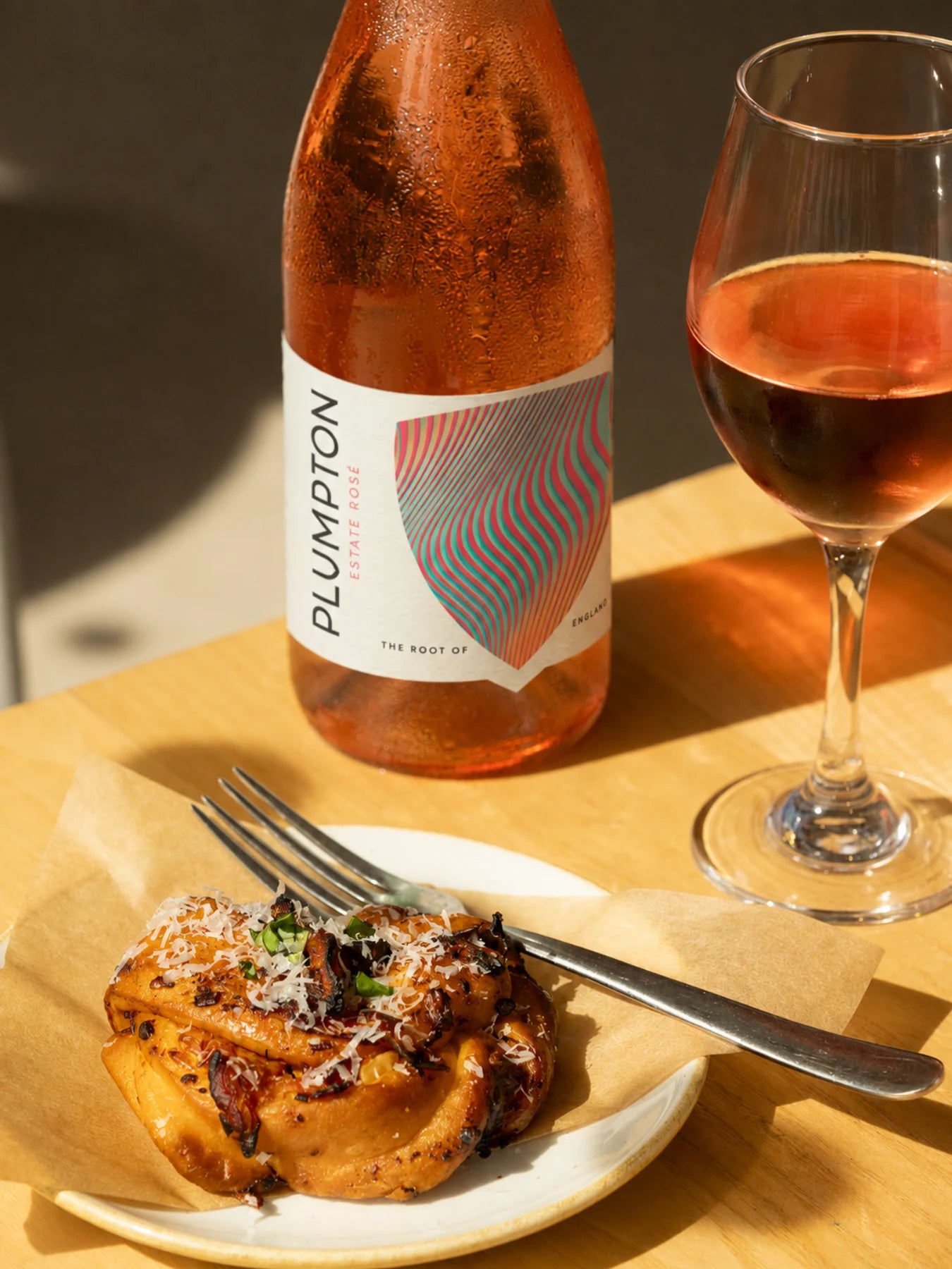 A bottle of Plumpton Estate Rose wine stands on a wooden table beside a filled wine glass, with a plate of pasta in the foreground on a warm, sunlit tabletop.