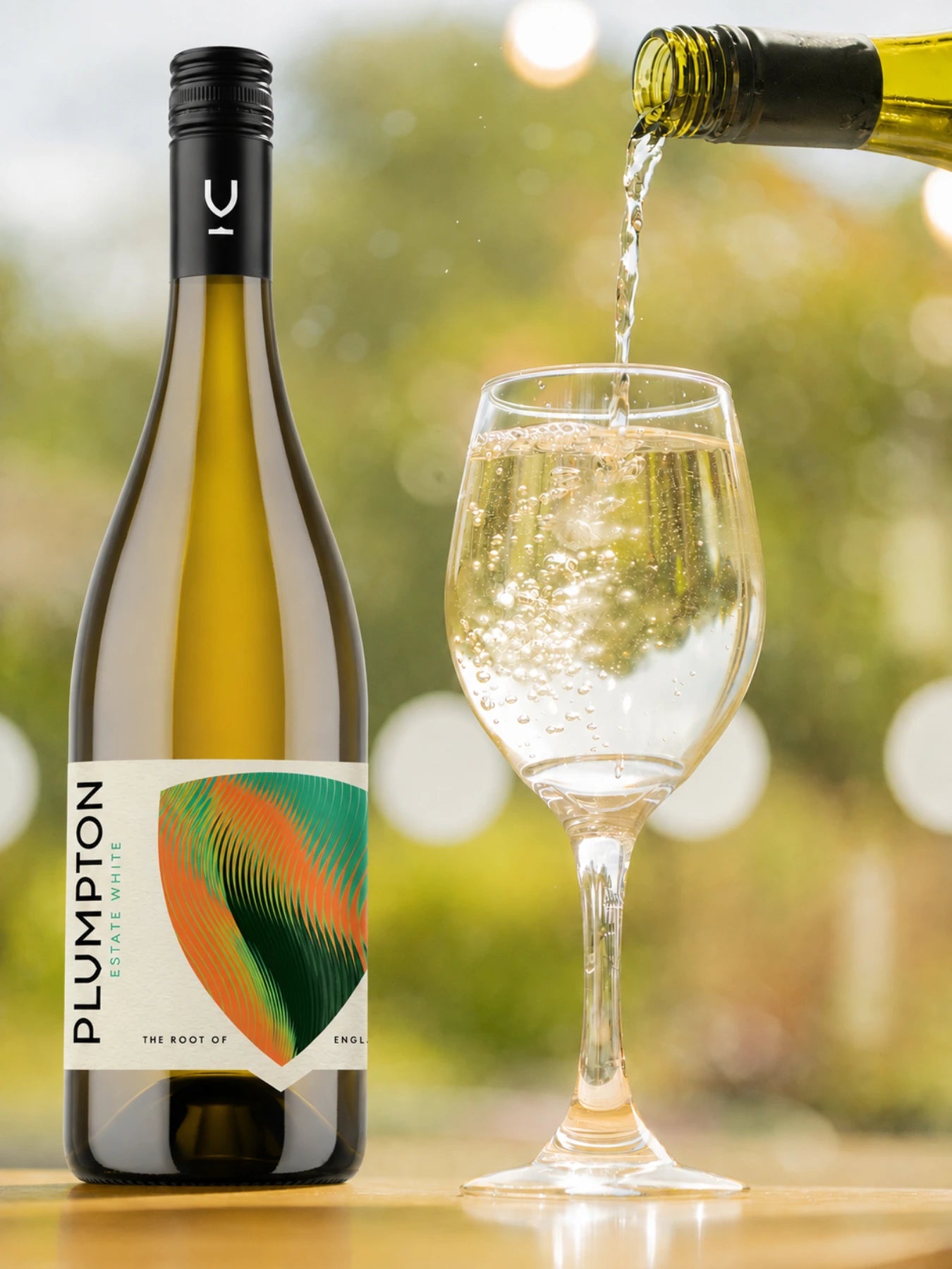 A bottle of Plumpton white wine stands on a wooden surface next to a wine glass, with white wine being poured into the glass. The background is a soft, blurred outdoor bokeh.