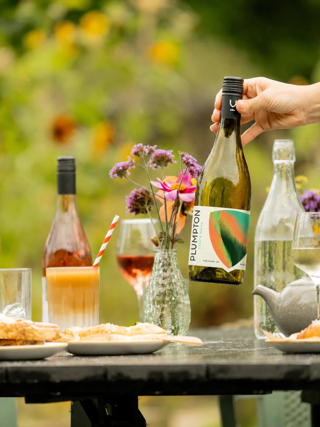 A hand holds a bottle of Plumpton rosé wine in an outdoor garden setting, with a table in the foreground holding plates, a drink with a striped straw, and flowers.